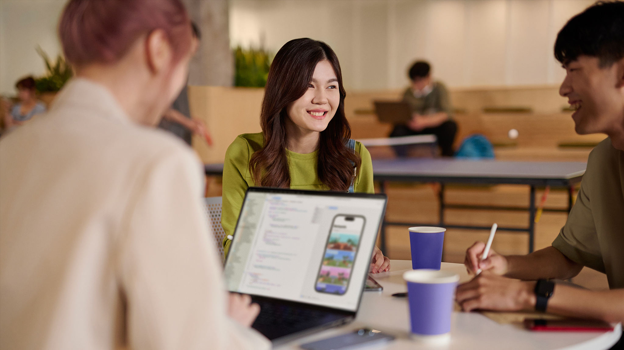 A woman in a green sweater sits at a table with two people, one of whom is working on an open laptop.