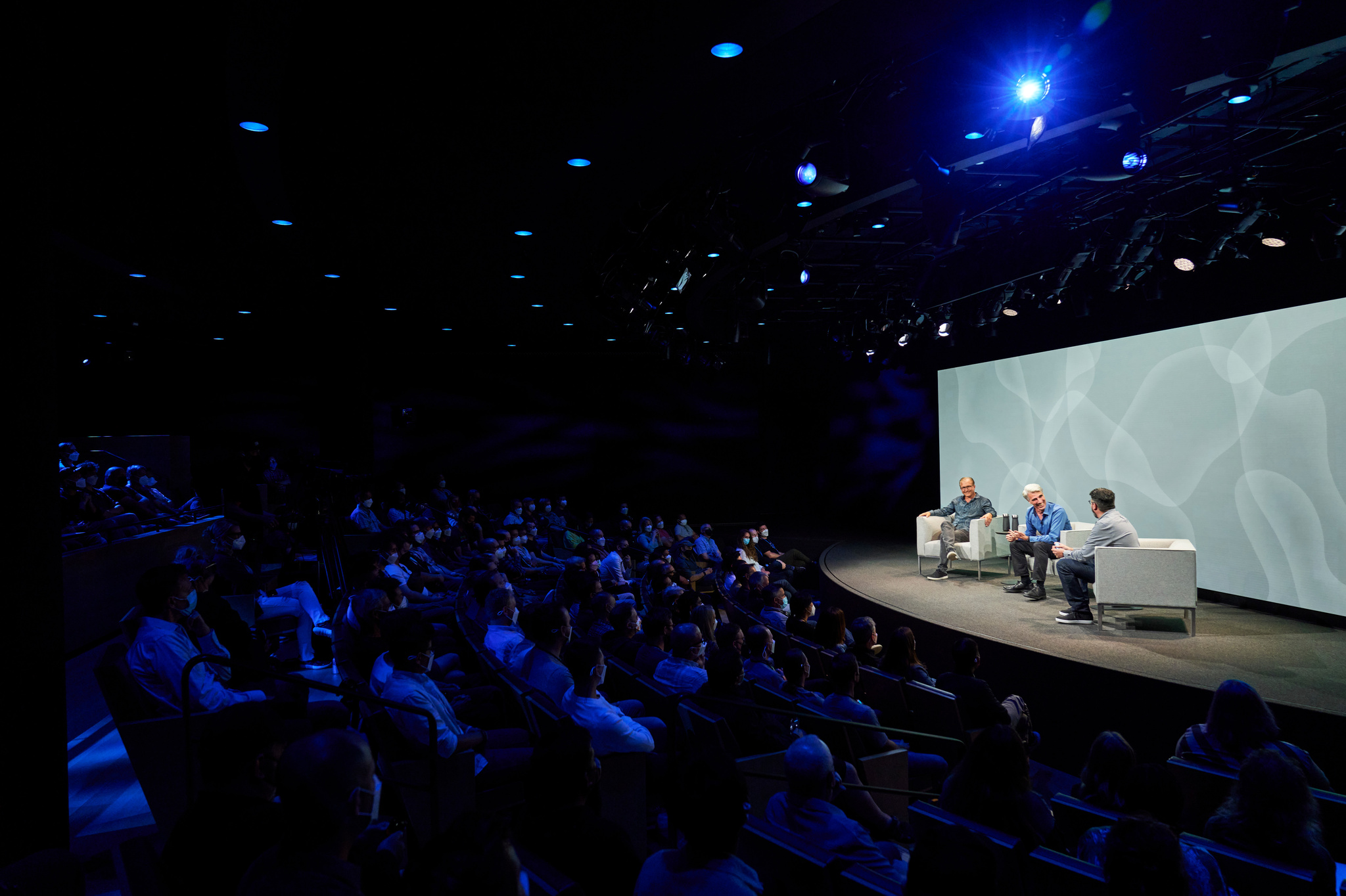 Photo of the Developer Center with dimmed lights and greg Joswiak, Craig Federighi, and John Gruber on stage