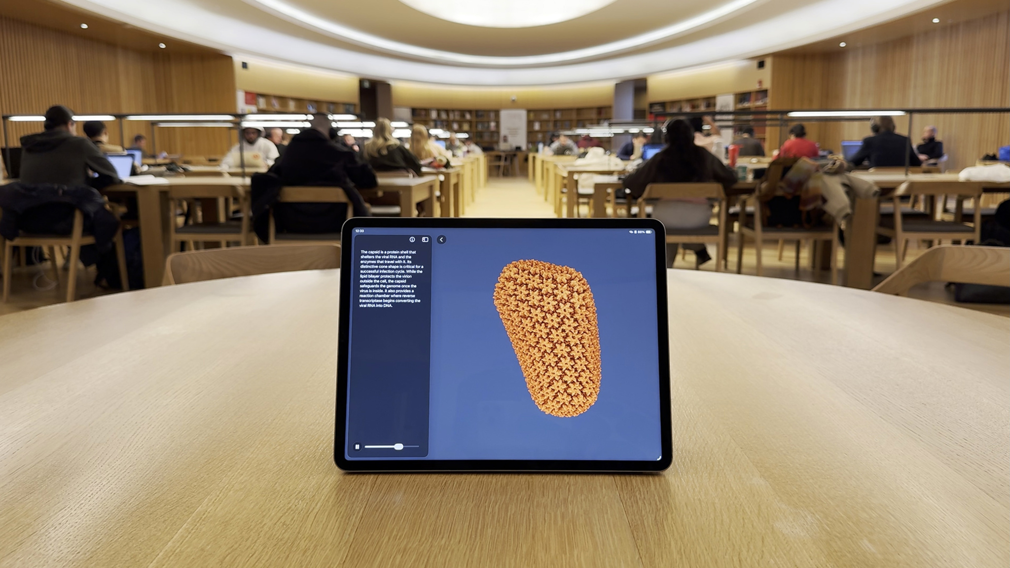 An iPad stands upright on a wooden table in a warmly lit library study hall, with rows of people working at long tables softly blurred in the background. The iPad screen displays a 3D molecular model of an orange, cone-shaped viral capsid against a deep blue background, accompanied by a panel of descriptive text on the left side of the screen. A playback scrubber at the bottom of the screen suggests the model is part of an interactive, guided experience.