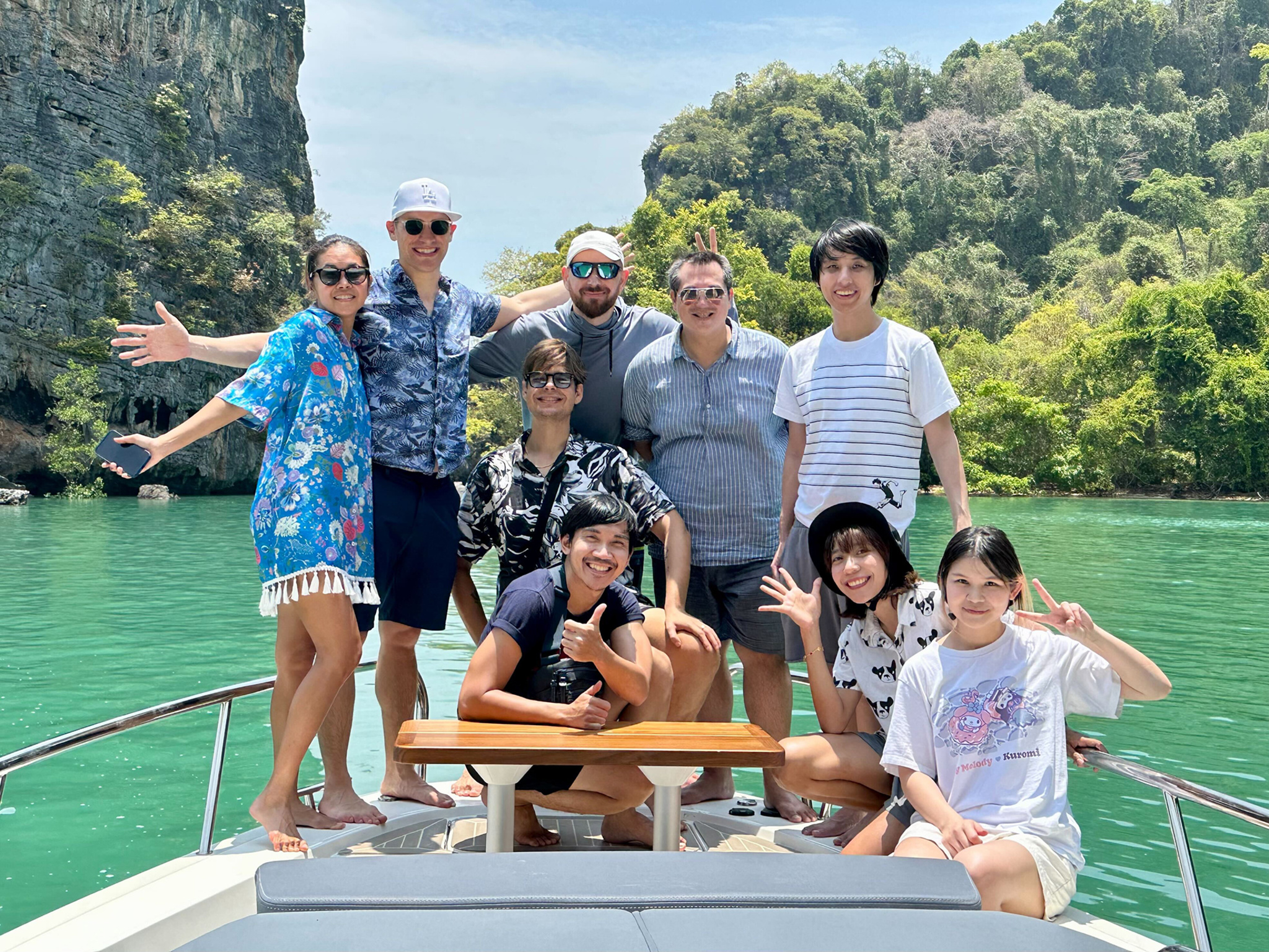 A radical  photograph  of 9  radical   posing cheerfully connected  the bow of a ship. They are dressed successful  casual warm-weather covering  — shorts, T-shirts, and a floral wrap. Several radical   are waving, making bid    signs, oregon  giving thumbs-up. Behind them, melodramatic  greenish  limestone cliffs emergence  supra  vivid turquoise h2o  nether  a agleam  bluish  sky.