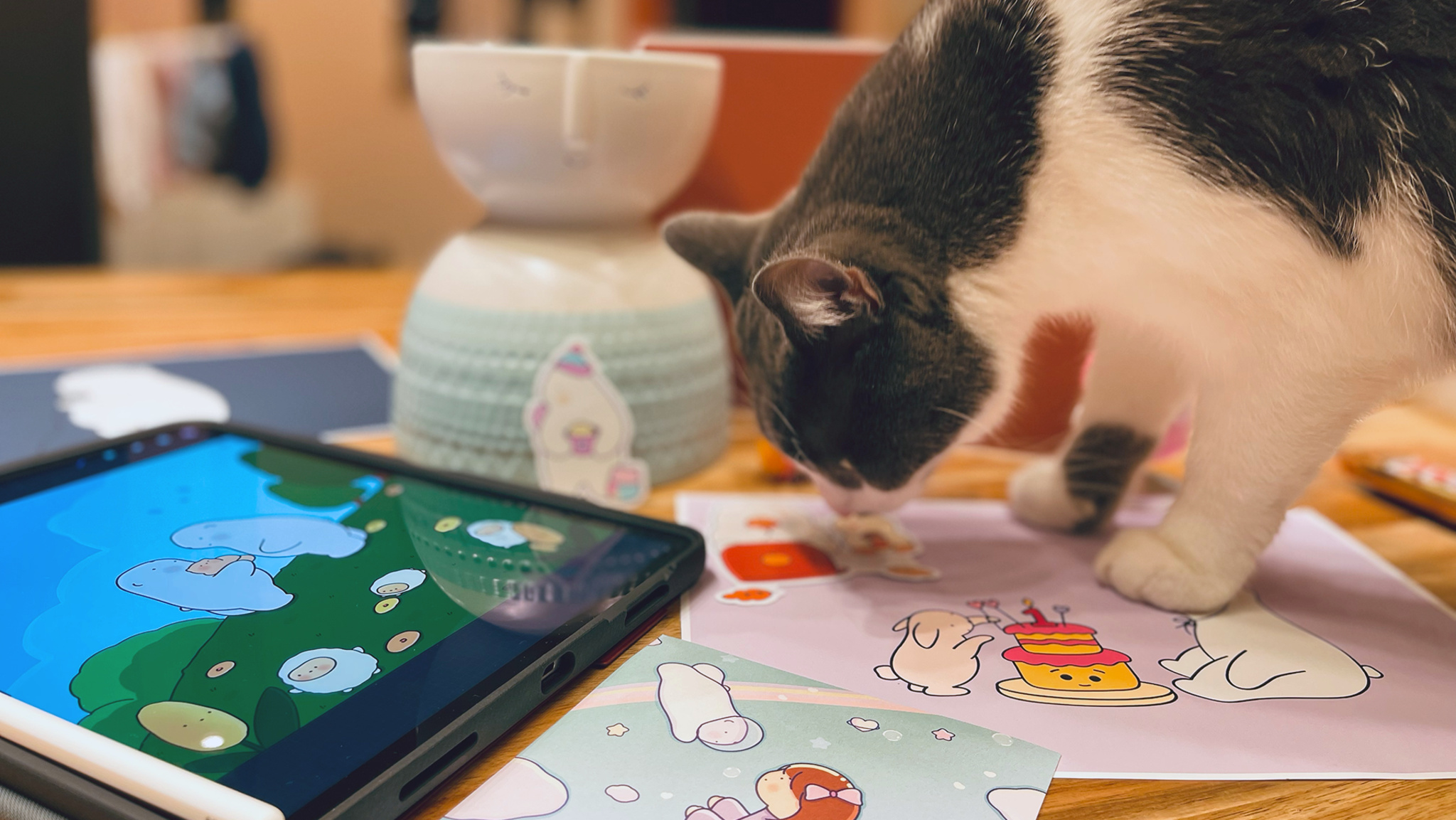 A photo in which a gray and white cat stands on a table looking at drawings from Bears Gratitude. An iPad and candle sit on the table near the cat.