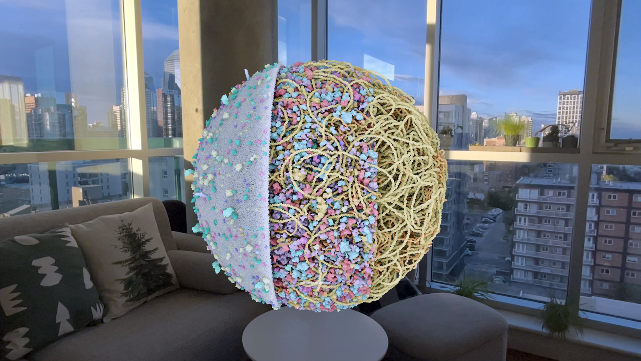 A cross-sectioned spherical 3D model of what appears to be a cell or organelle hovers above a small side table in a living room, rendered in {Apple Vision Pro} passthrough. The model’s interior reveals a dense tangle of yellow filaments studded with pastel pink, teal, and lavender molecular clusters. A city skyline stretches across large windows in the background, and a couch with patterned throw pillows is visible to the left.