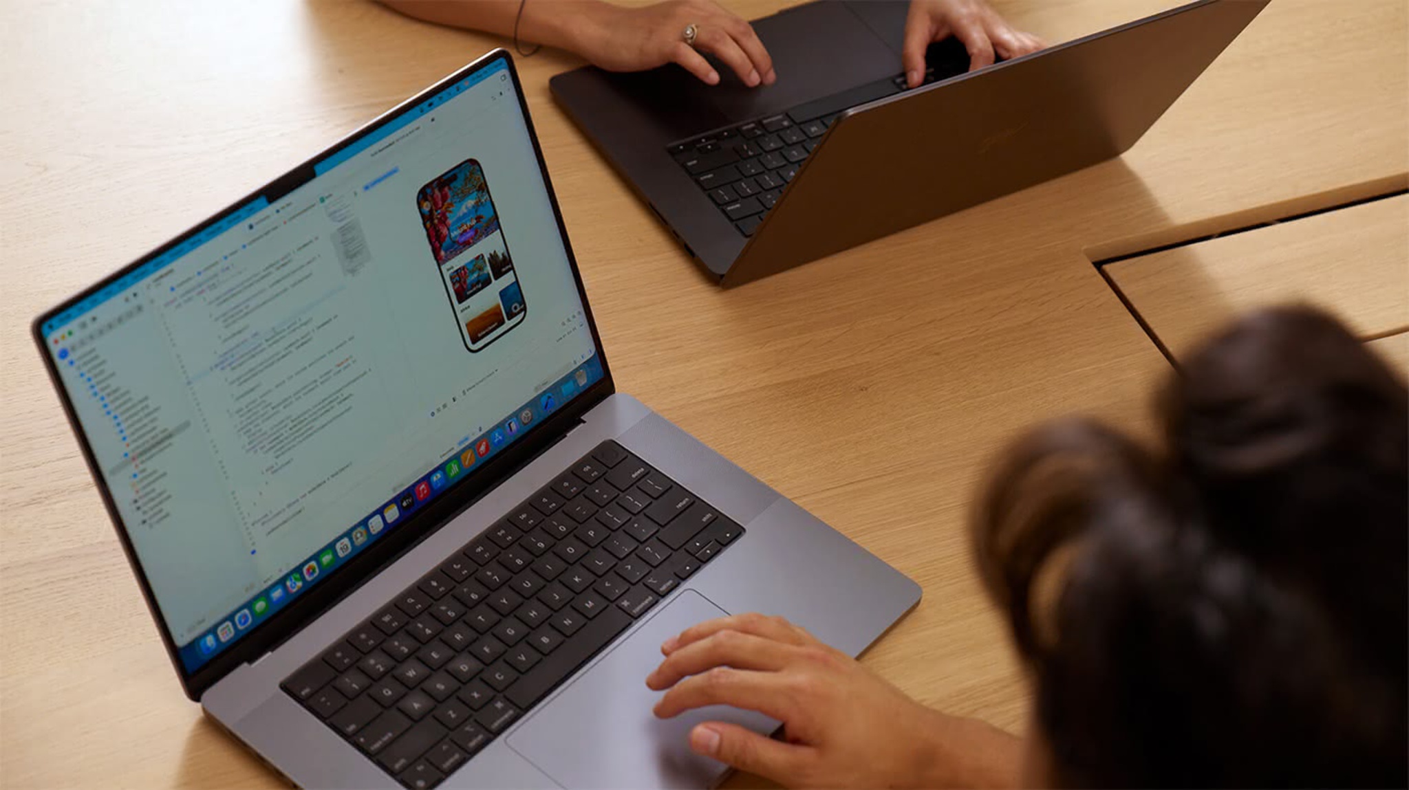 An overhead view of two people working side by side at a light wood table, each on a MacBook Pro. The foreground laptop displays Xcode with an iOS app simulator showing a colorful travel or photo-browsing app on a virtual iPhone screen. The second person’s hands are visible in the upper portion of the frame, typing on their MacBook, suggesting a collaborative development or code review session.