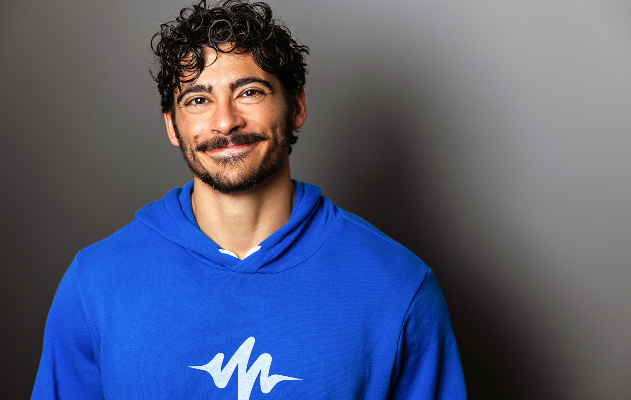 A photo of Speechify founder Cliff Weitzman, who is wearing a blue hoodie and standing against a gray wall.