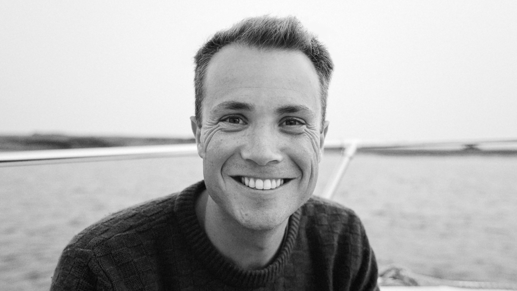 A black and white image of Tucker MacDonald, who is wearing a dark sweater and sitting on a boat on the water.