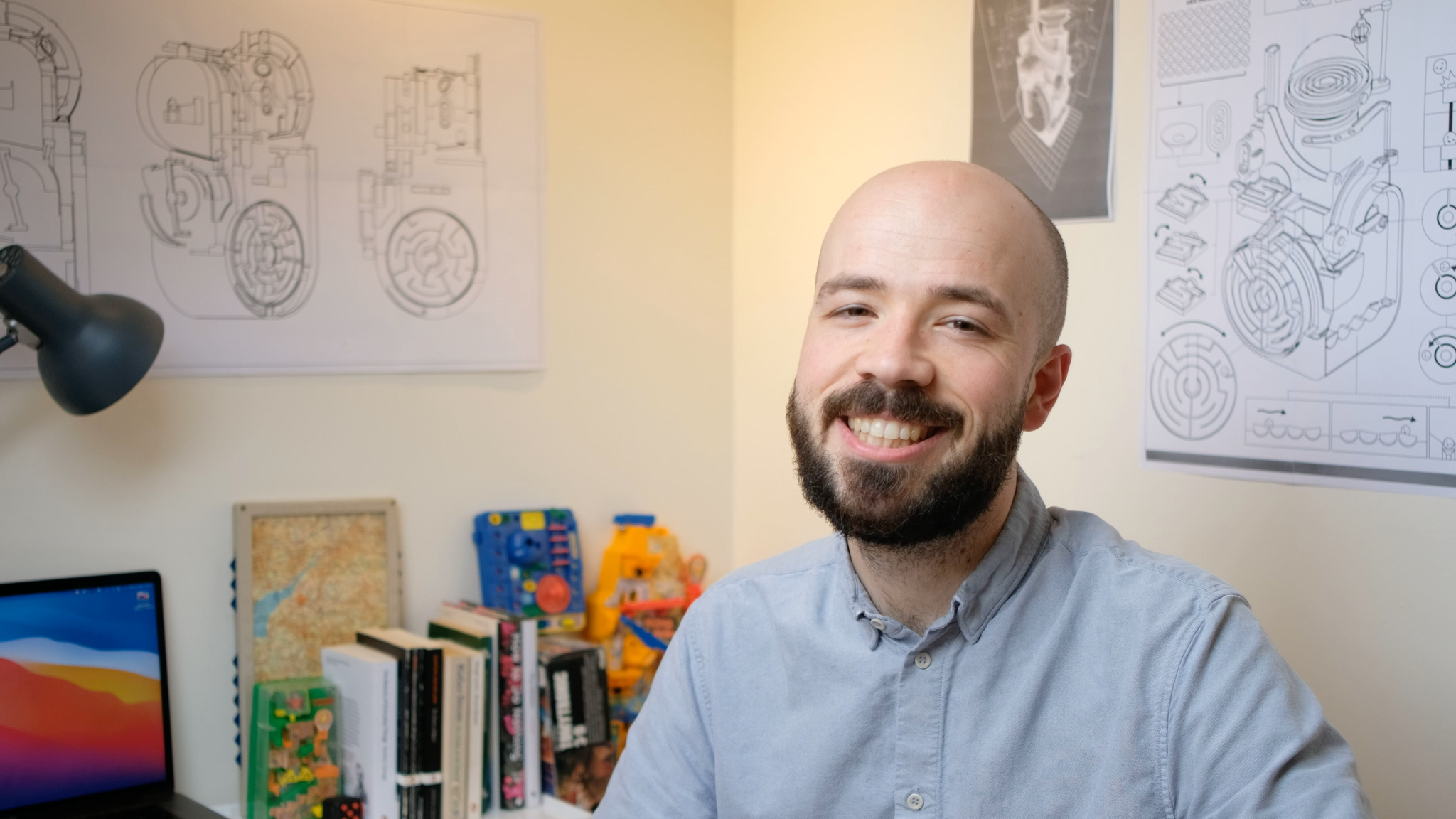 A portrait of Automatoys developer Steffan Glynn seated in his home office, where black and white sketches of the game’s levels hang on the wall around him.