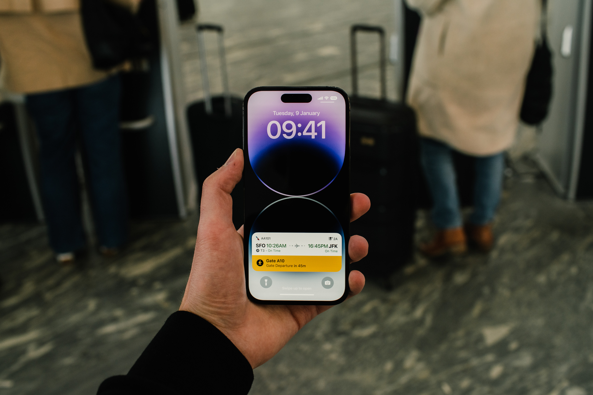 A photo of a hand holding an iPhone showing the app Flighty, which is displaying flight information in a Live Activity format.