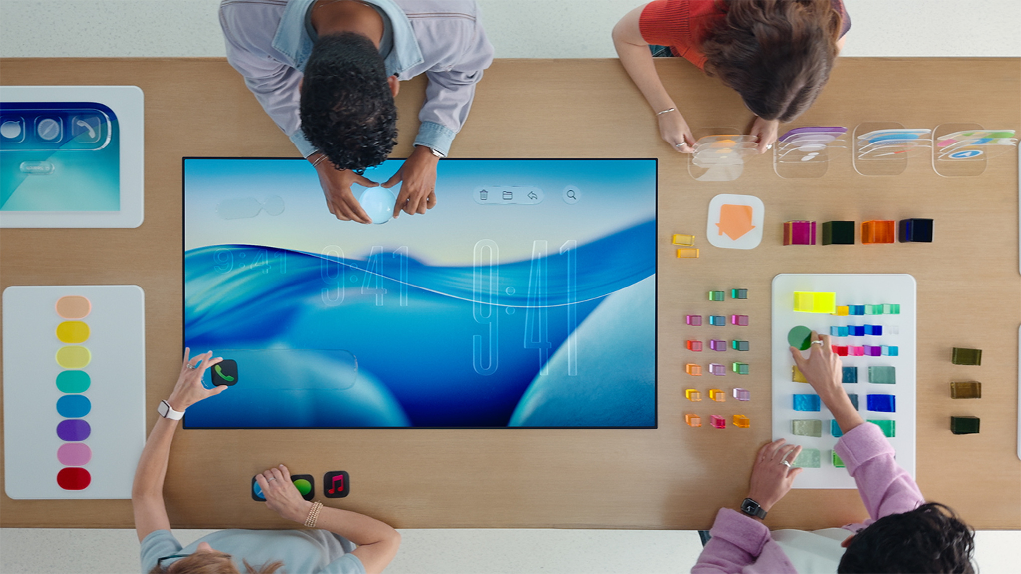 A top-down view of several people working at a table assembling colorful objects that evoke the new design and Liquid Glass.