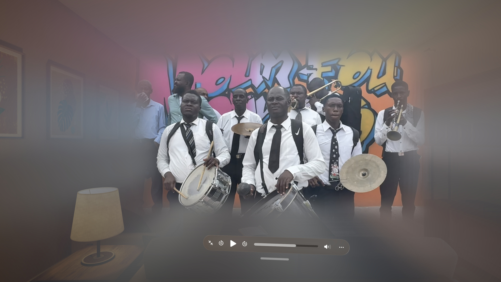 A brass and percussion band of approximately eight musicians performs in front of a vibrant graffiti mural in orange, pink, and blue. The musicians in the foreground wear white dress shirts, dark ties, and suspenders, and are playing snare drums and cymbals, while brass players with a trombone and trumpet stand behind them. The image is a paused video frame, shown with a media player control bar visible along the bottom.