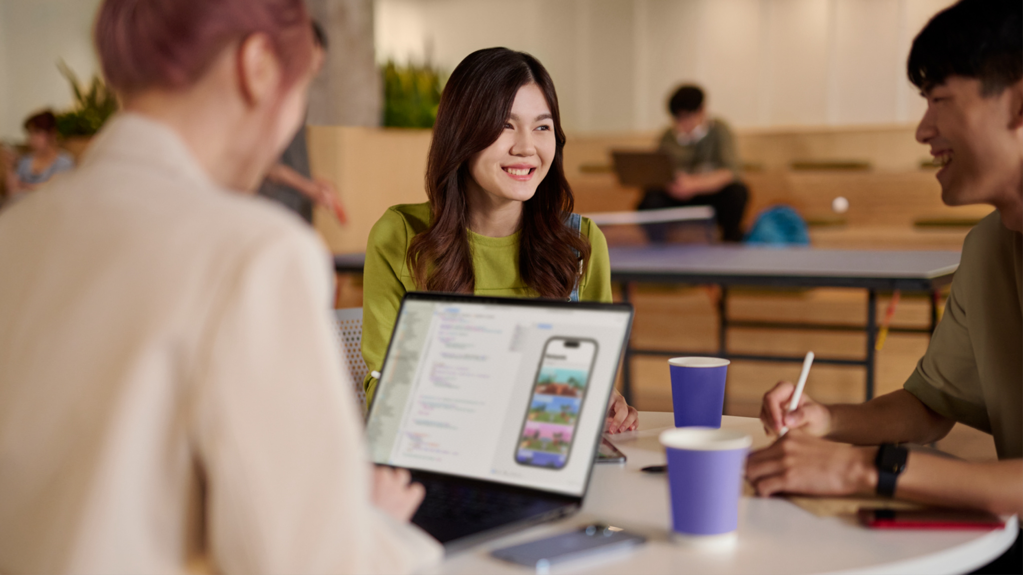 A woman in a green sweater sits at a table with two people, one of whom is working on an open laptop.