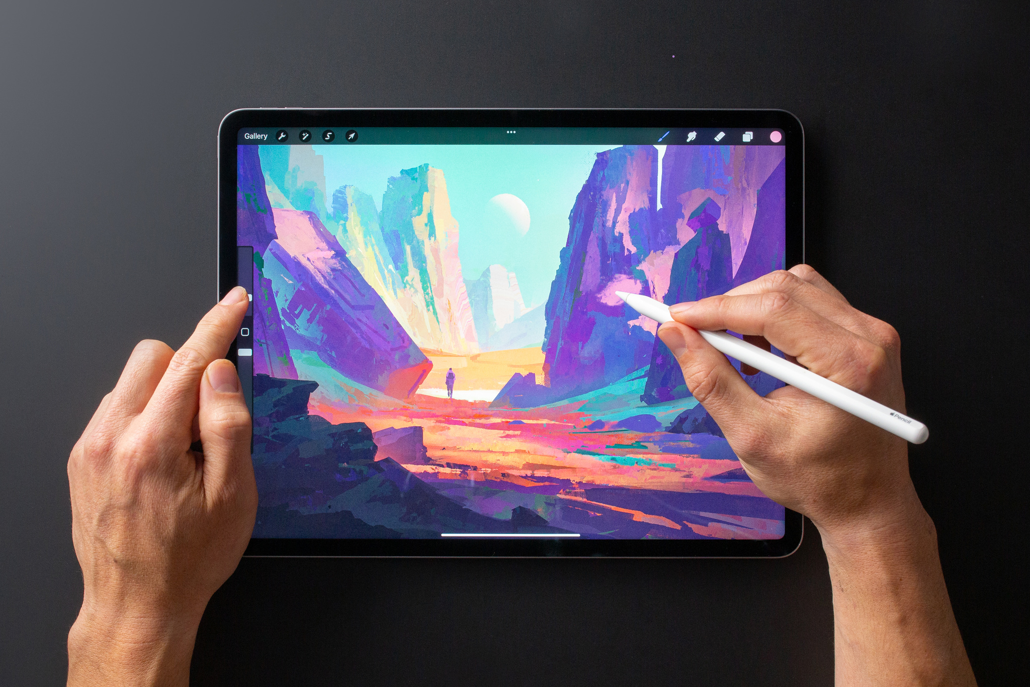 An image of the app Procreate on an iPad. A person uses their left hand to manipulate sliders on the left side of the screen, and their right hand to hover Apple Pencil over the abstract, colorful canvas.