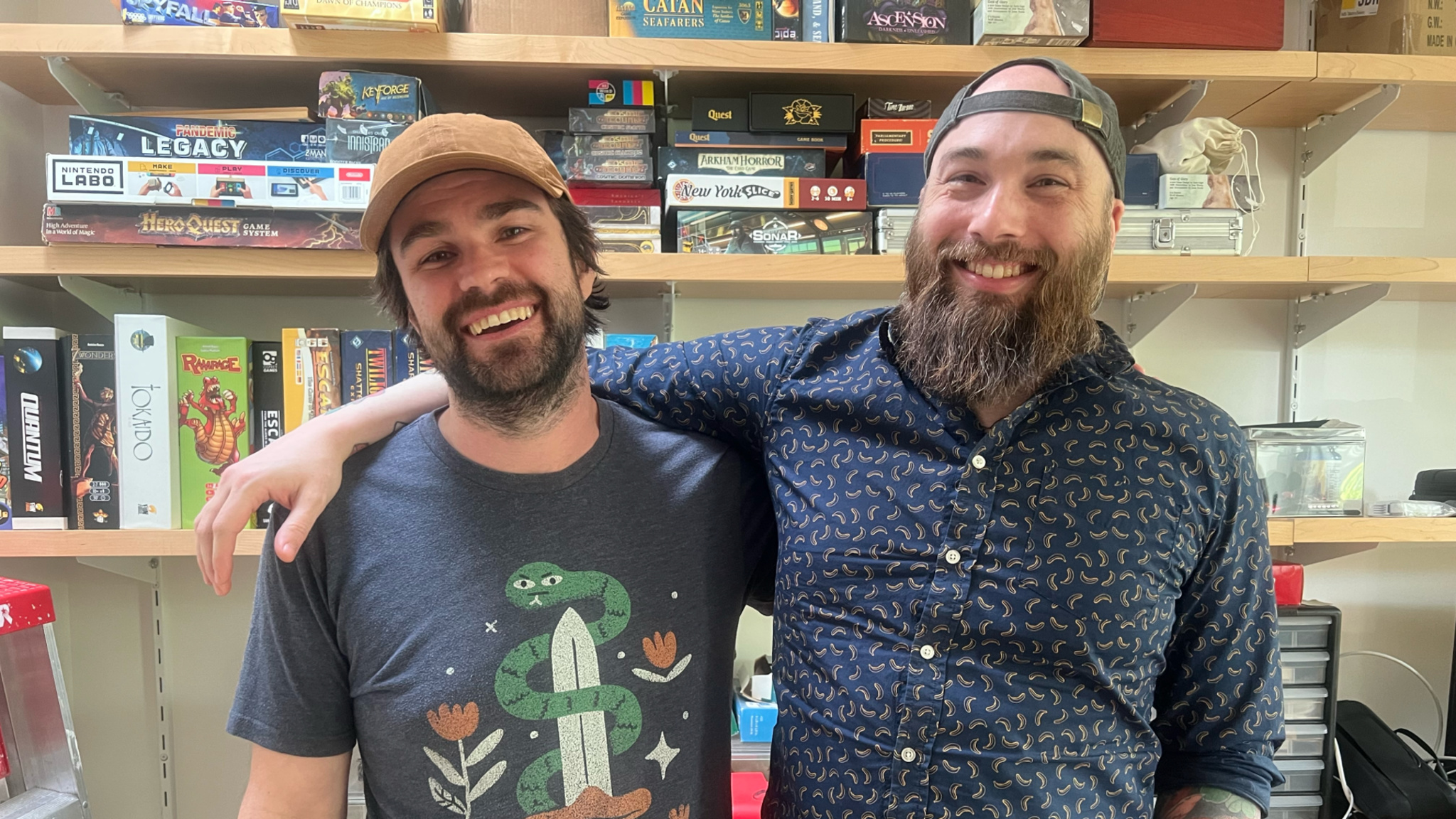 Zach Gage, left, and Jack Schlesinger stand in their studio in front of a series of shelves that hold several board games.