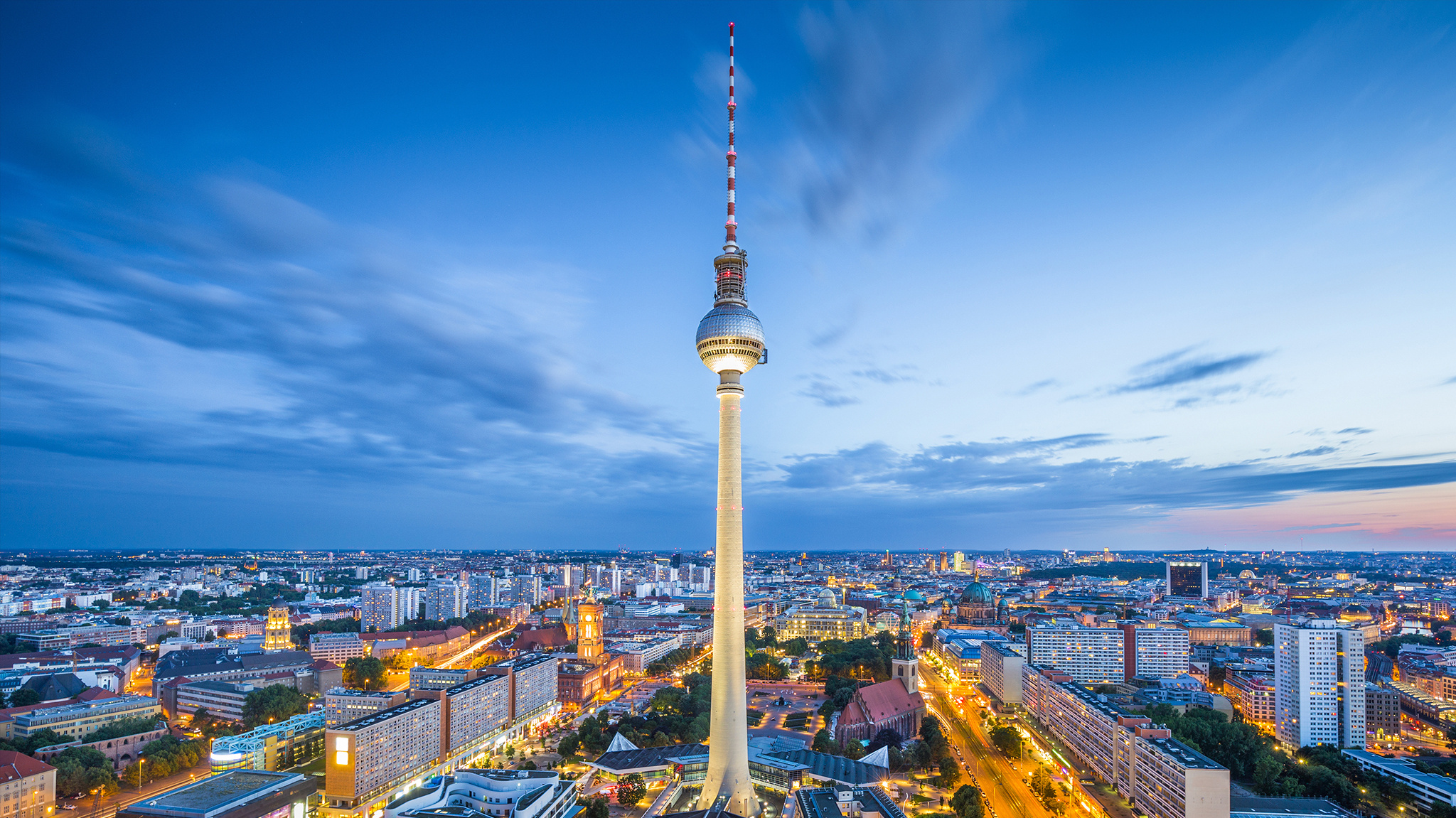 Aerial view of Berlin at dusk, with the Fernsehturm television tower rising prominently above the illuminated city skyline against a blue evening sky.
