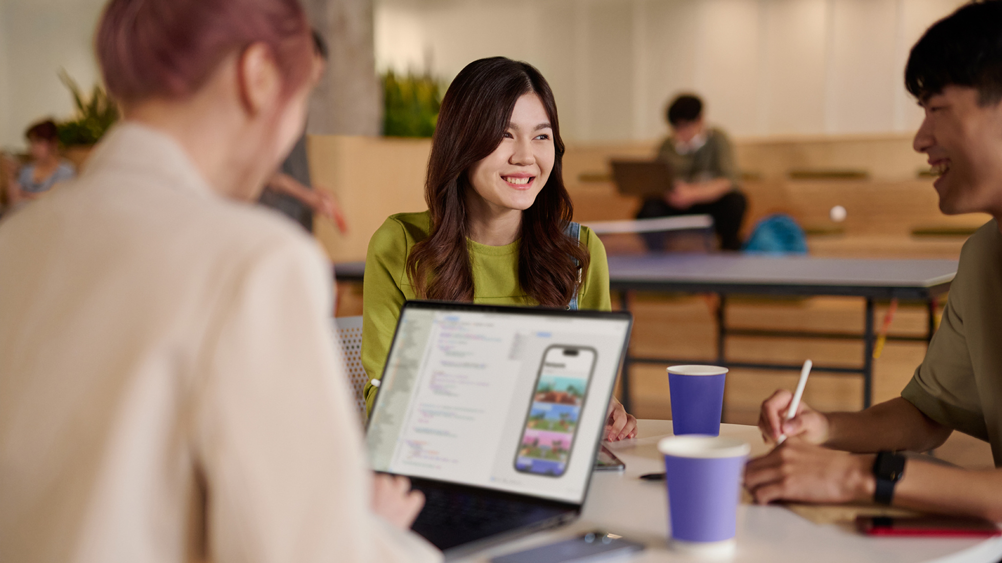 A woman in a green sweater sits at a table with two people, one of whom is working on an open laptop.